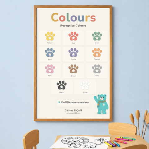 Landing Ready® Colours Wall Chart displayed in a modern family living room.A framed 'Colours Recognise Colours' educational poster hanging on a light blue wall above a children's drawing table with crayons and wooden tools. The poster features colorful animal paw prints and text including 'Yellow', 'Red', 'Green', 'Blue', 'Purple', 'Orange', 'Pink', 'Brown', 'Gray', 'Black', and 'White', with a small blue bear illustration and the brand name 'Canvas & Quill'. 